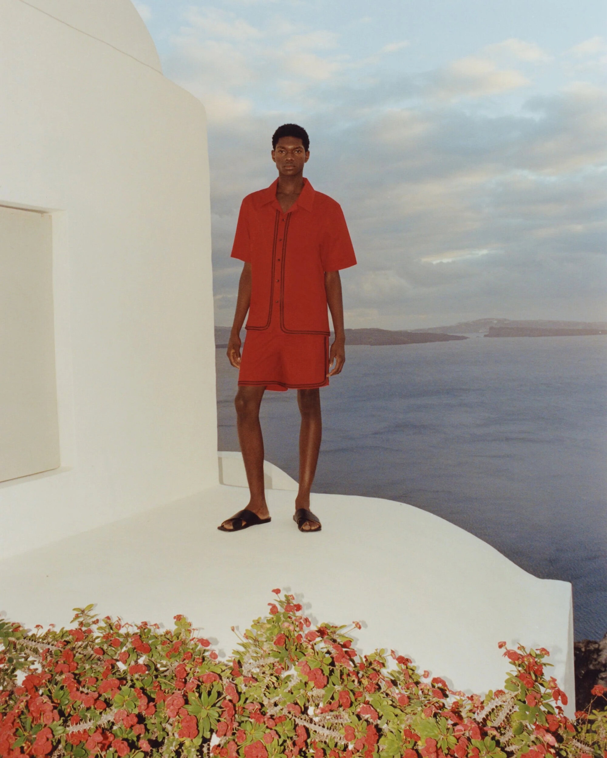Man in red shirt and shorts stands on a white balcony overlooking the sea with red flowers in the foreground under a cloudy sky.