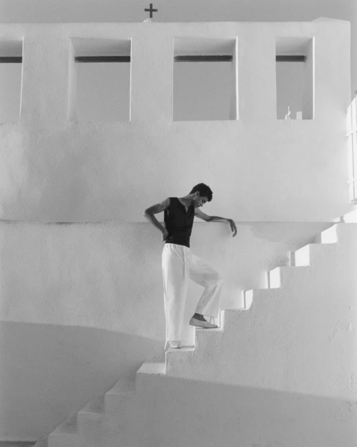 A man in a tank top and white pants ascends a white staircase indoors, with natural light and a cross above a window in the background.