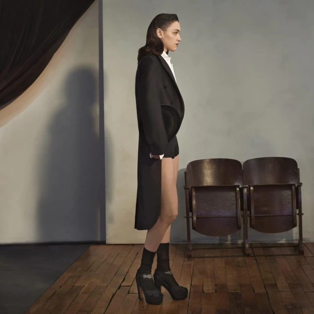 Woman in profile wearing black coat, shorts, and high heels in studio with theatrical chairs and soft lighting, for fashion editorial.