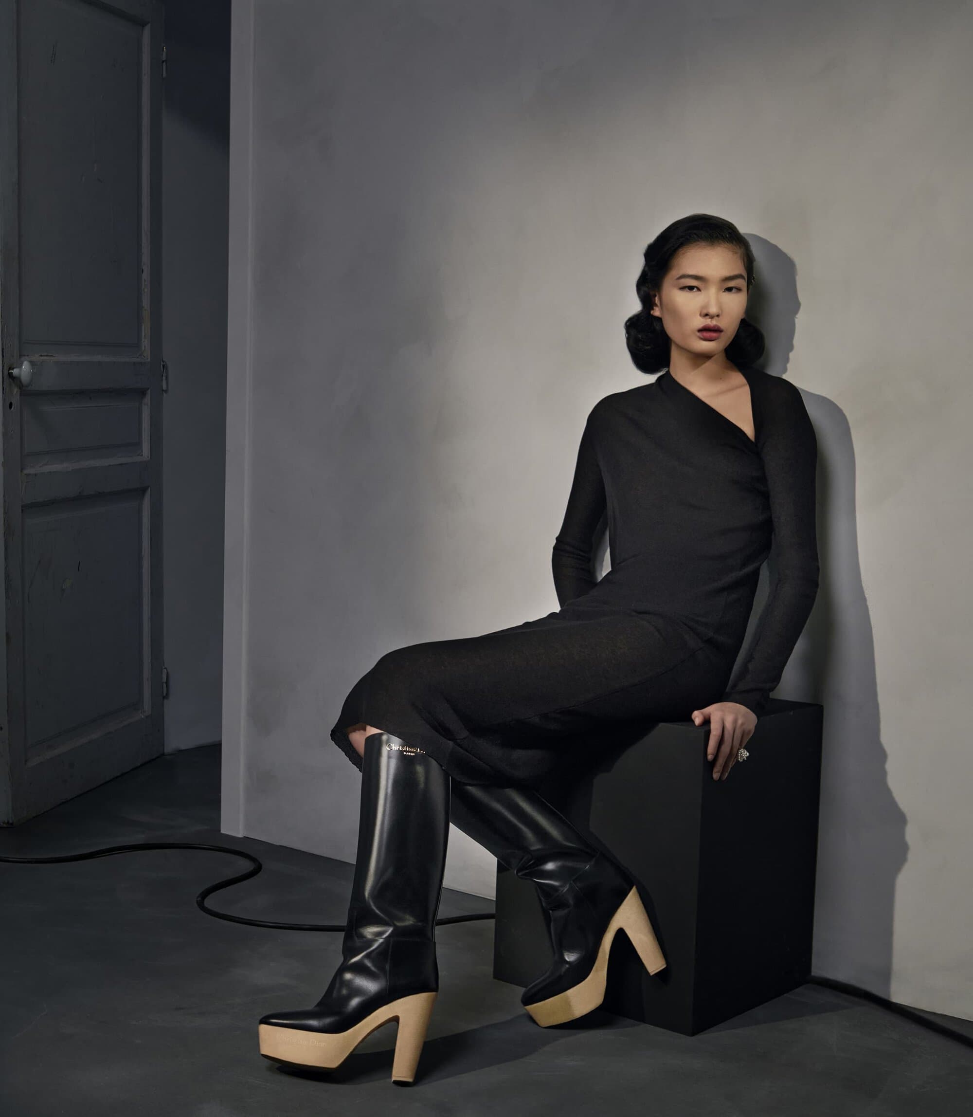 A woman in a black dress and platform boots sits on a black cube against a neutral wall near a closed door, posing in a fashion editorial.