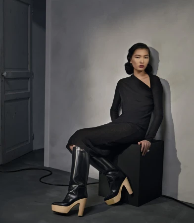 A woman in a black dress and platform boots sits on a black cube against a neutral wall near a closed door, posing in a fashion editorial.