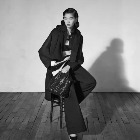 Model seated on a stool wearing a dark coat and trousers holding a structured handbag against a plain background.
