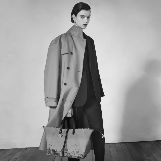 Monochrome portrait of a woman in a trench coat and suit holding a printed handbag, with a clean backdrop and studio lighting.
