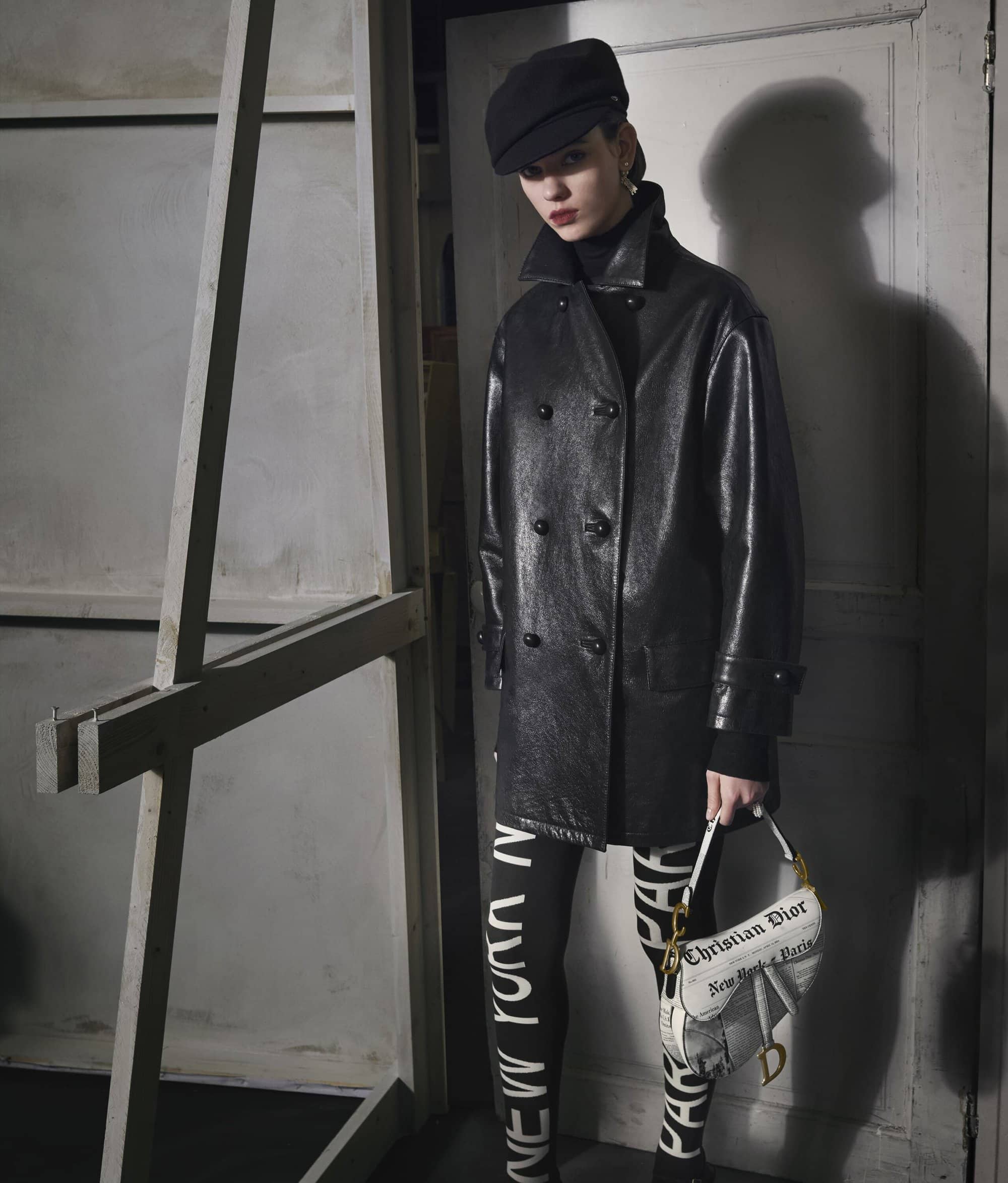 Model in black leather coat and cap holding printed saddle bag against studio wall