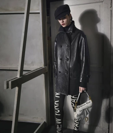 Model in black leather coat and cap holding printed saddle bag against studio wall