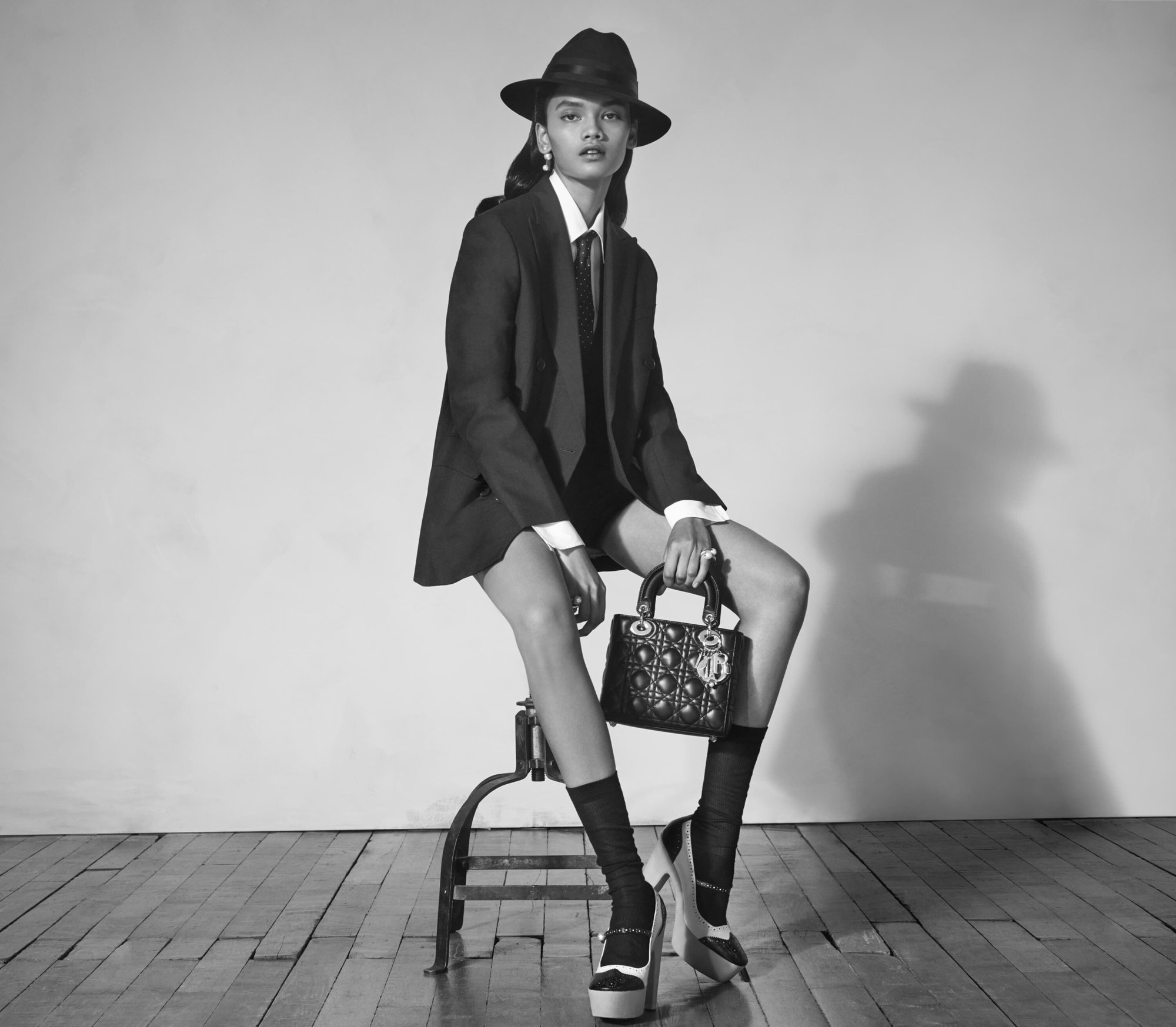 Model wearing dark suit jacket, fedora hat, and tie seated on a stool holding a quilted handbag against a plain studio background.