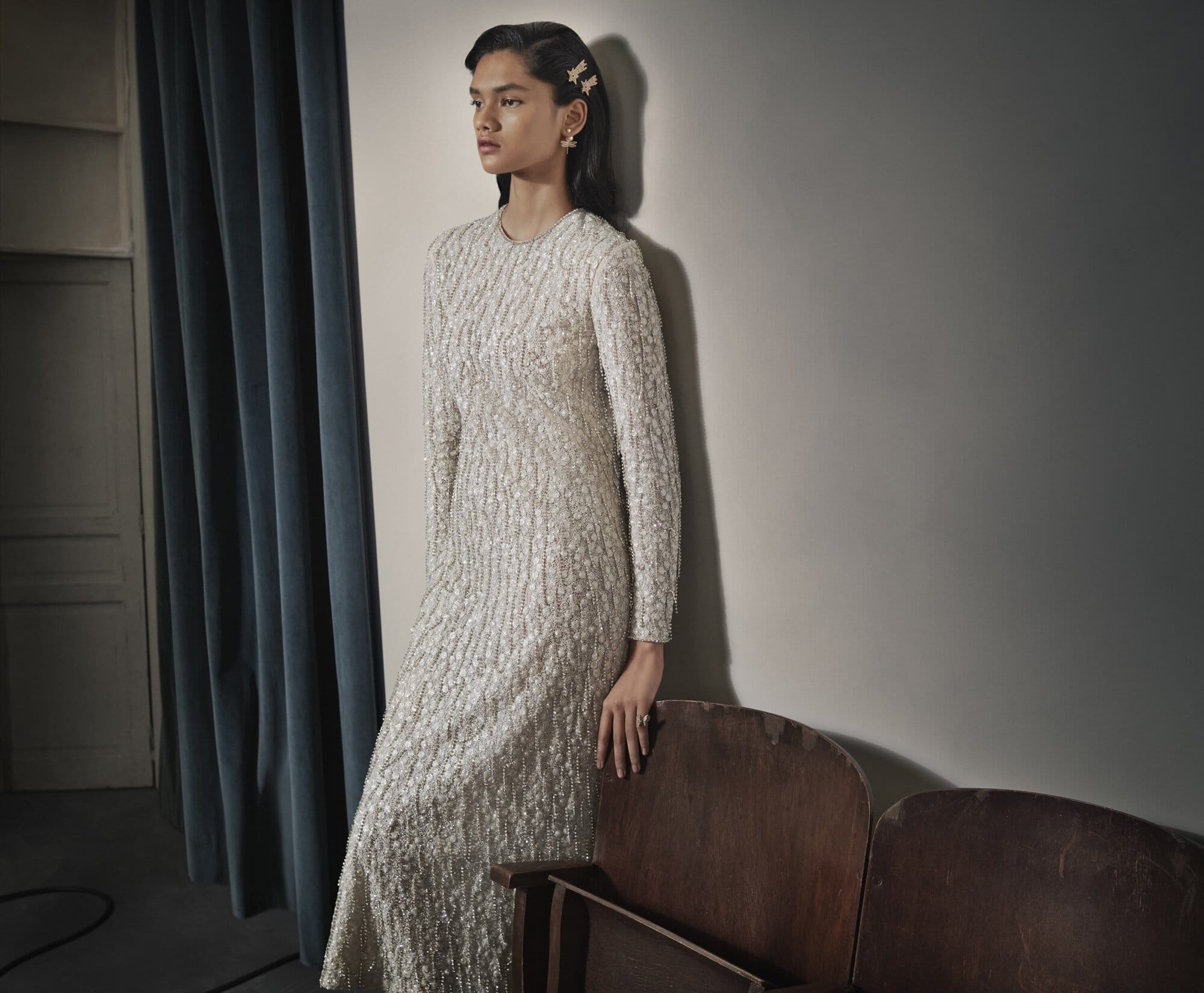 Elegant woman in a long-sleeved, textured white dress leans against a wall with vintage theater seats. The lighting is soft and neutral.