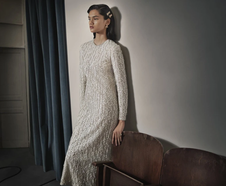 Elegant woman in a long-sleeved, textured white dress leans against a wall with vintage theater seats. The lighting is soft and neutral.