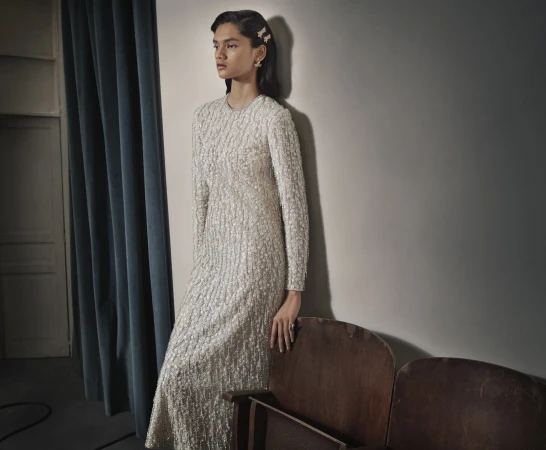 Elegant woman in a long-sleeved, textured white dress leans against a wall with vintage theater seats. The lighting is soft and neutral.