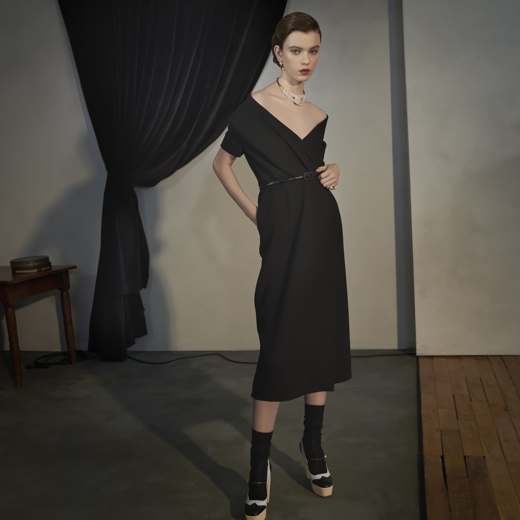 A stylish woman poses in a black dress, accessorized with a pearl necklace and belt, complemented by black socks and platform heels.