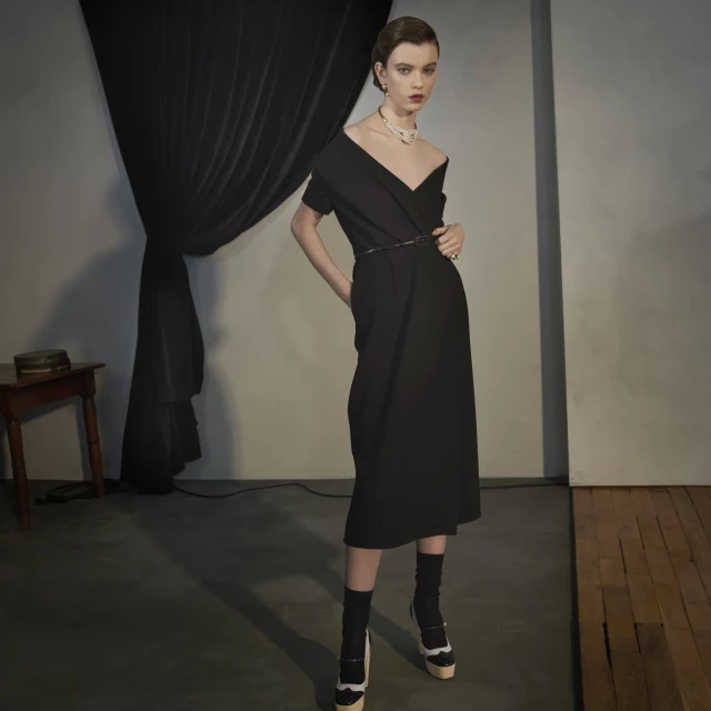A stylish woman poses in a black dress, accessorized with a pearl necklace and belt, complemented by black socks and platform heels.