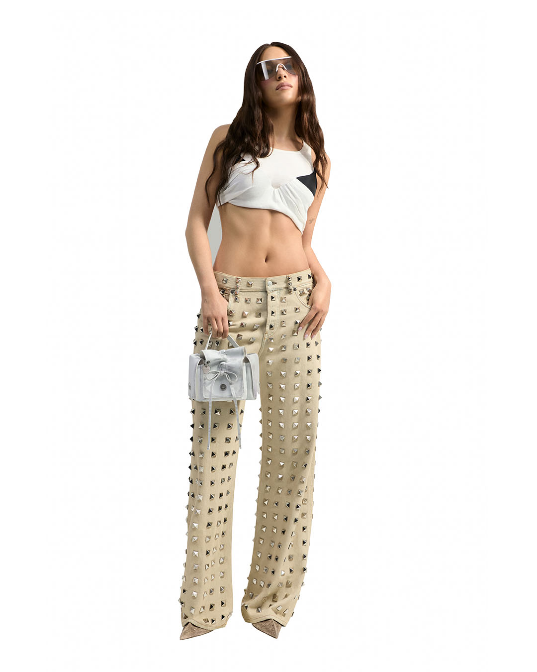 A fashion model wearing a white halter top and studded pants, poses with a small, silver handbag against a clean white background.