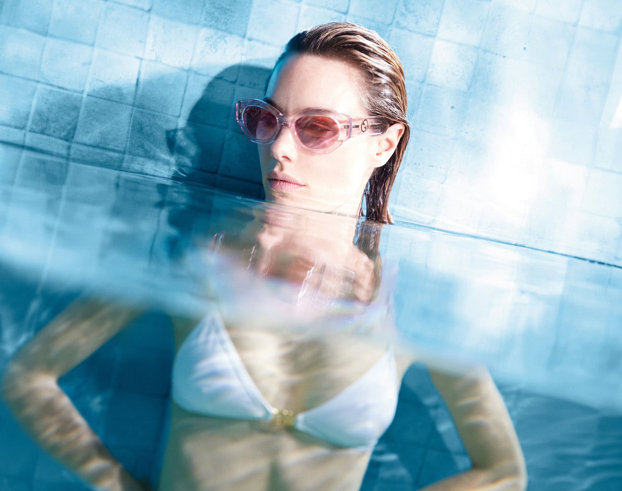 Blonde woman in sunglasses and white bikini partially submerged in pool, turquoise tile background. Wet hair and neutral expression.