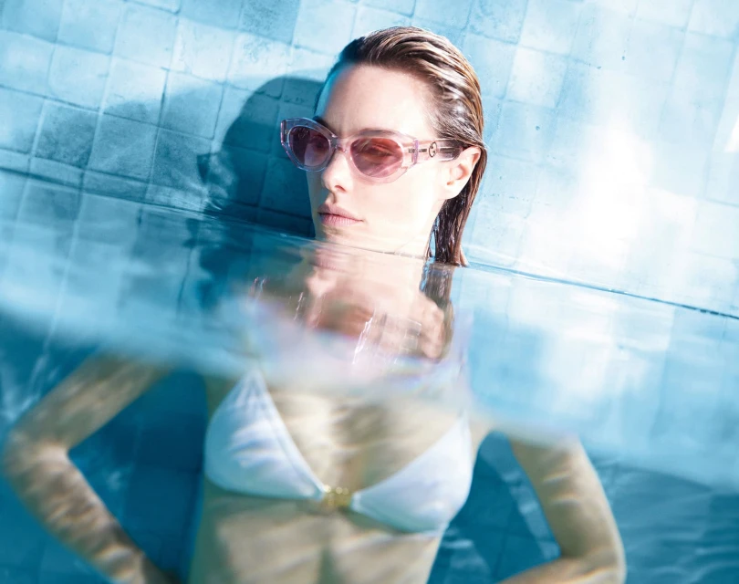 Blonde woman in sunglasses and white bikini partially submerged in pool, turquoise tile background. Wet hair and neutral expression.