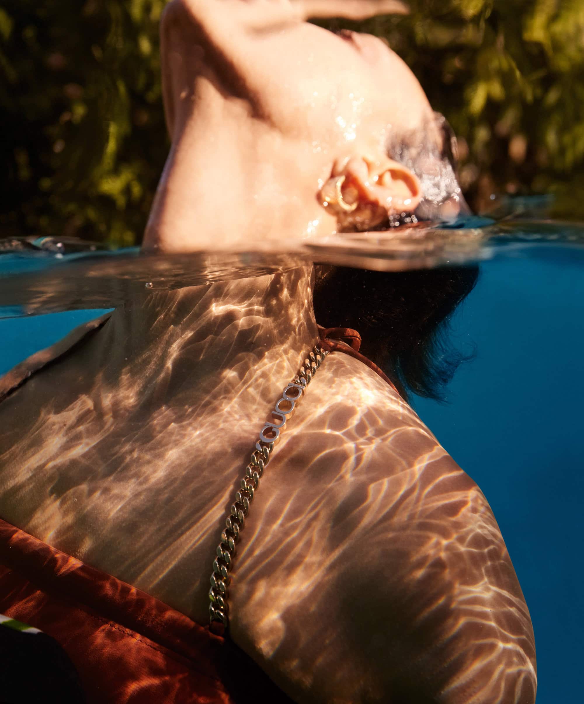 Adult female in pool with filtered sunlight on skin. Gold chain necklace visible above water. Lush green plants in background.