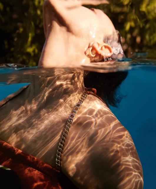 Adult female in pool with filtered sunlight on skin. Gold chain necklace visible above water. Lush green plants in background.