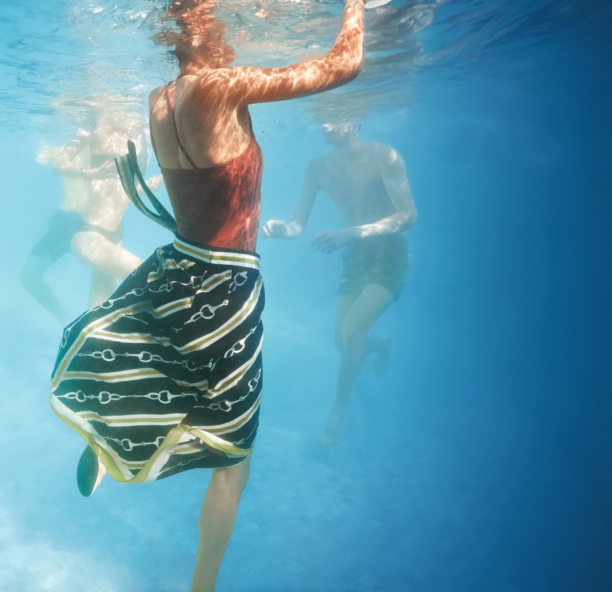 Underwater shot of a woman in a skirt and top, with two people swimming in the blurred background in blue water.
