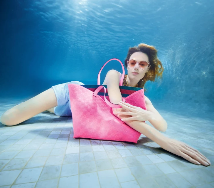 Underwater shot of a woman posing with a pink handbag in a swimming pool, creating a surreal and fashionable visual composition.