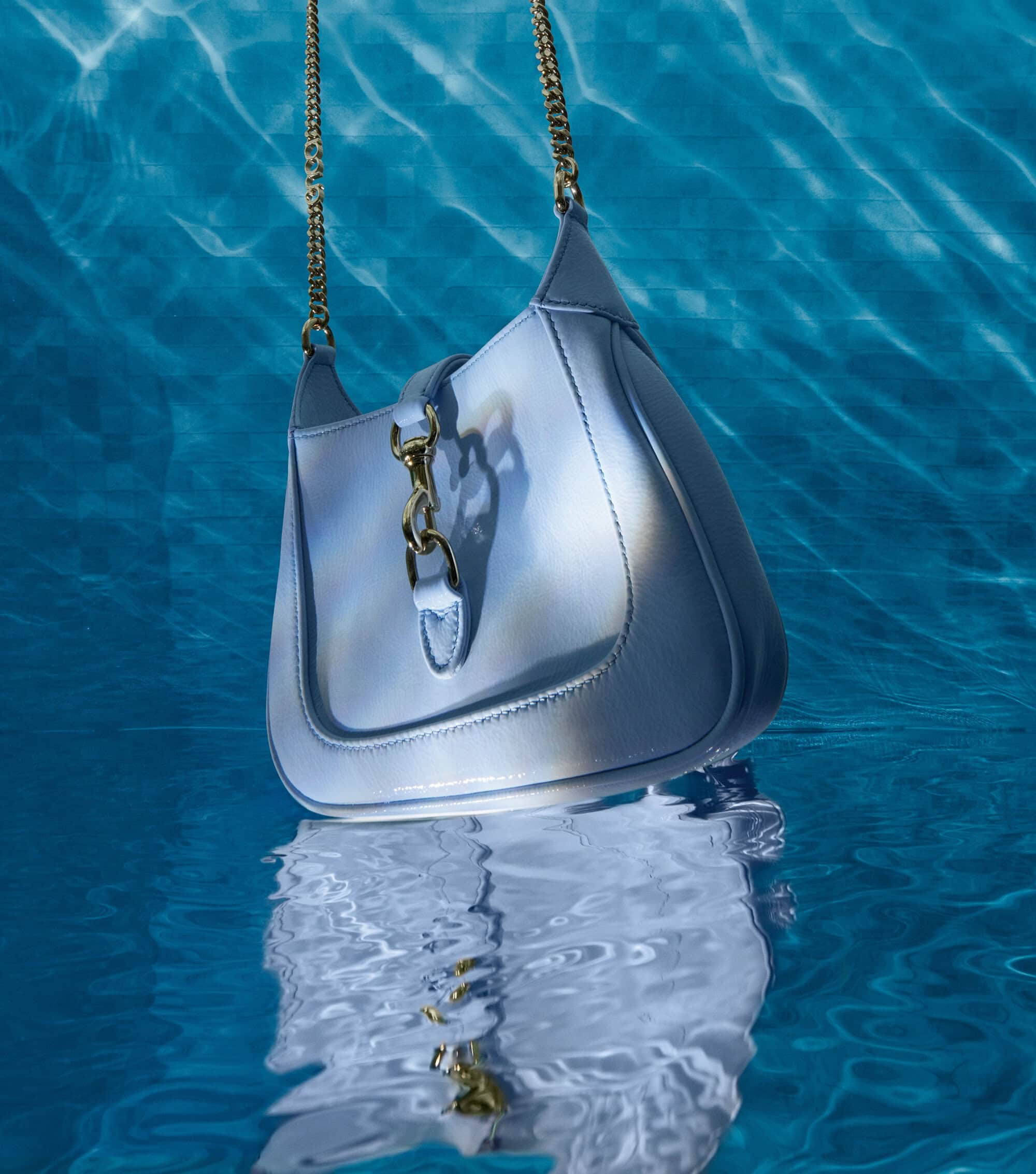 Pale blue leather purse with gold chain strap and hardware floating beneath the surface of clear blue swimming pool water.