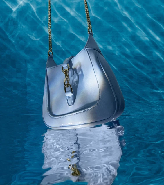 Pale blue leather purse with gold chain strap and hardware floating beneath the surface of clear blue swimming pool water.