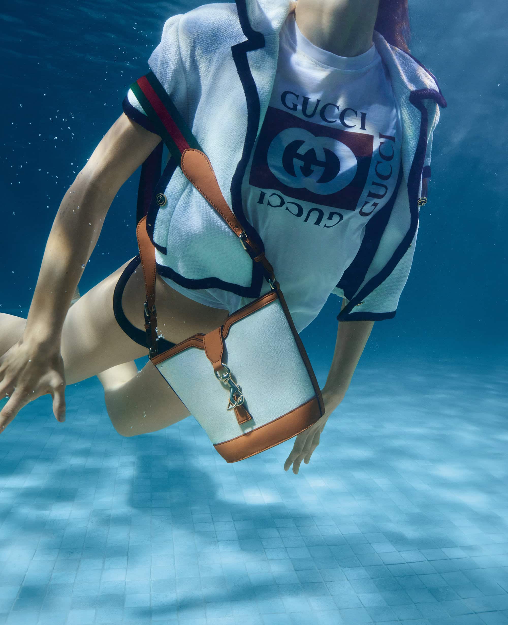 Fashion shot of a woman underwater in a pool, wearing a white Gucci t-shirt, jacket, and holding a white and brown purse.