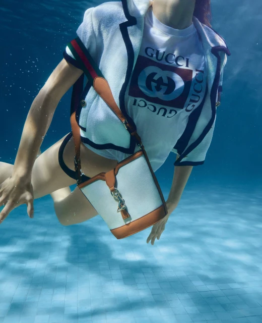 Fashion shot of a woman underwater in a pool, wearing a white Gucci t-shirt, jacket, and holding a white and brown purse.