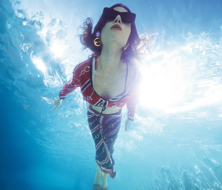 Woman with sunglasses poses underwater in a red and blue patterned outfit in clear blue water with sun rays and golden earrings.