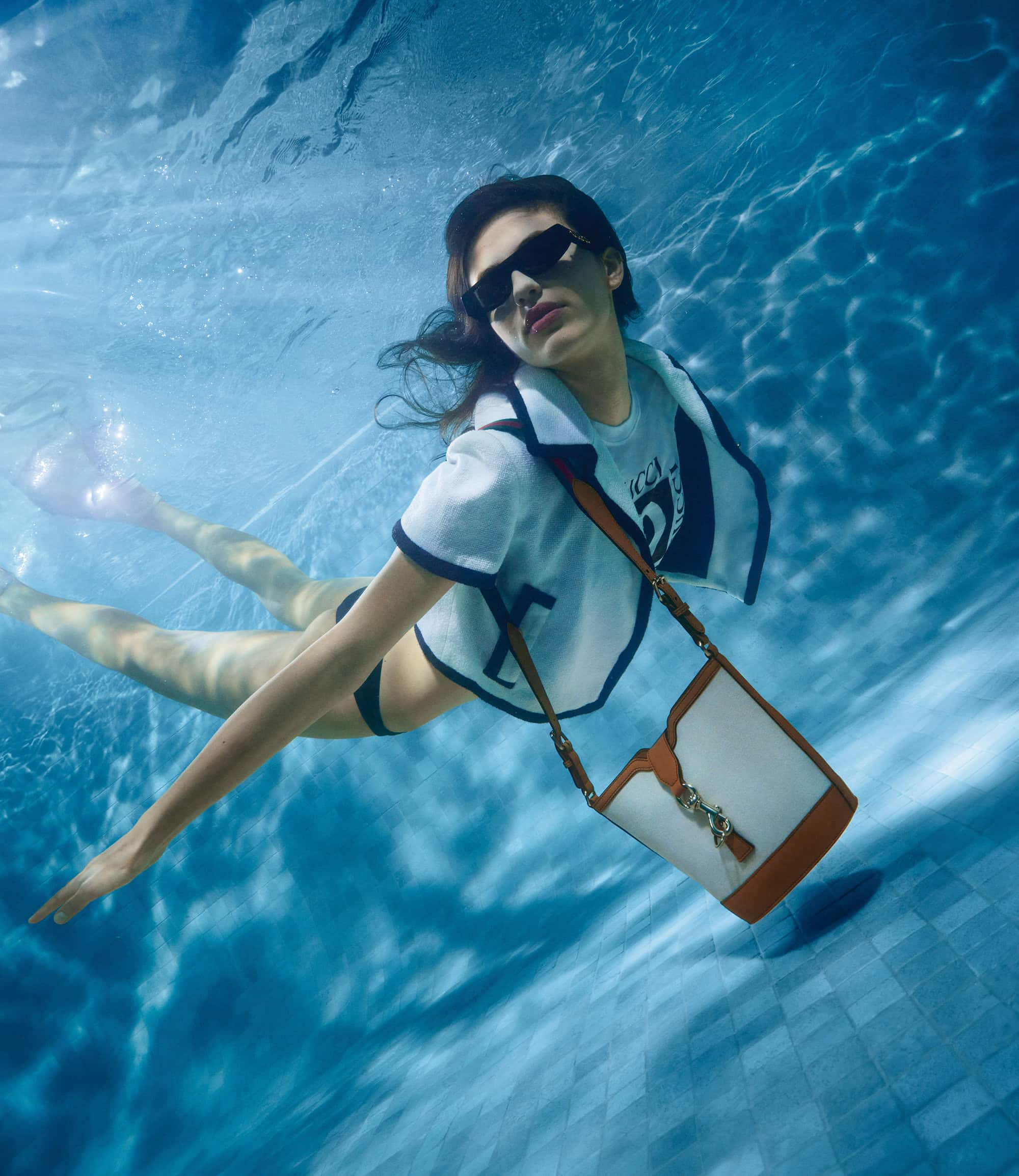 Woman swims underwater in a pool wearing sunglasses, cropped short sleeve shirt, bikini bottom, and carrying a white and brown purse.