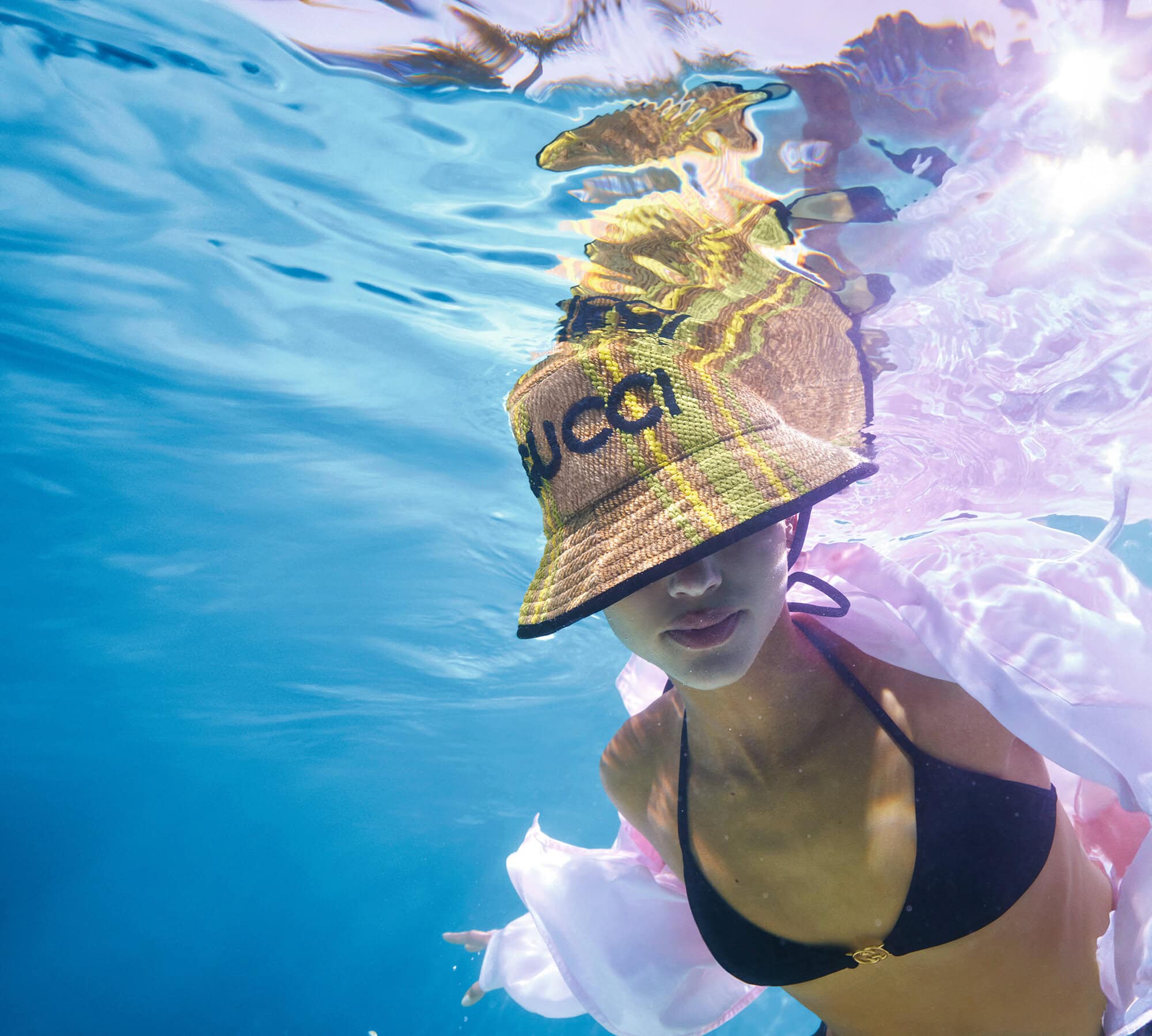 Woman swimming underwater in a black bikini and a patterned Gucci bucket hat, with sunlight filtering through the clear blue water.