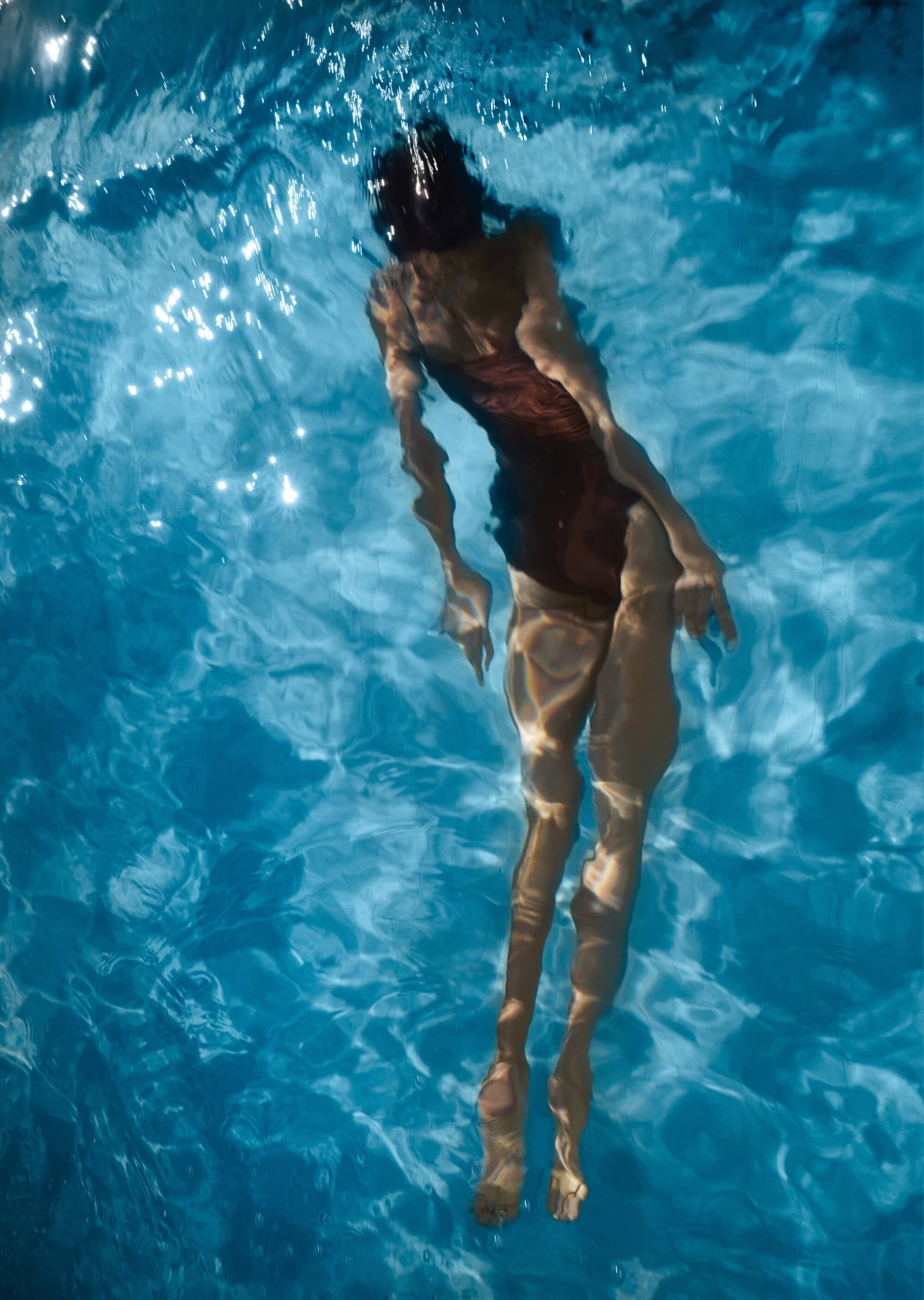 Woman in a maroon one-piece swimsuit swims underwater in a pool, filtered sunlight creating patterns on the pool's surface.