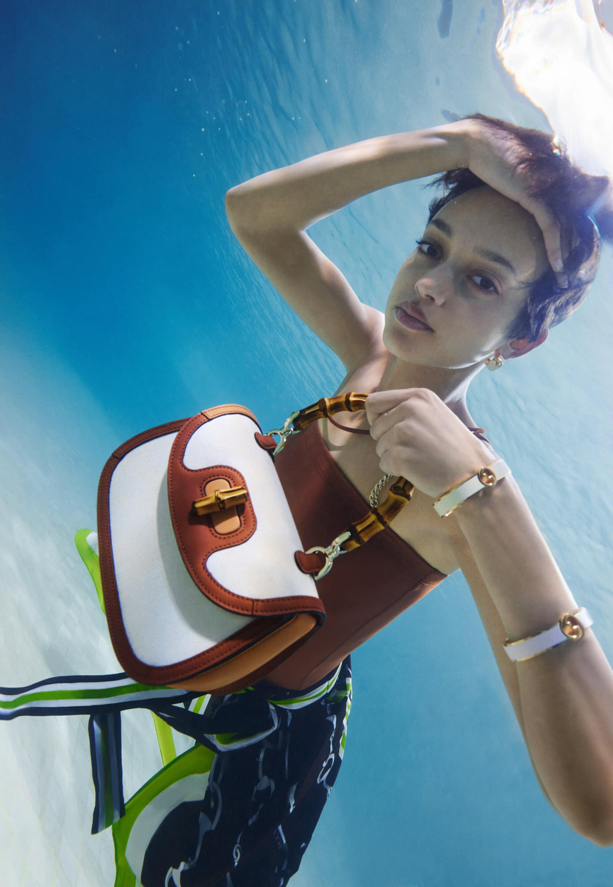 A woman underwater, wearing a brown top and a handbag, poses with her hand behind her head, while wearing bracelets.