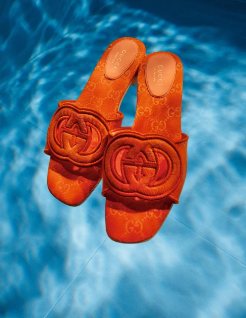Elevated shot of orange Gucci slides with a logo pattern and emblem, suspended over a rippled blue water surface creating light reflections.