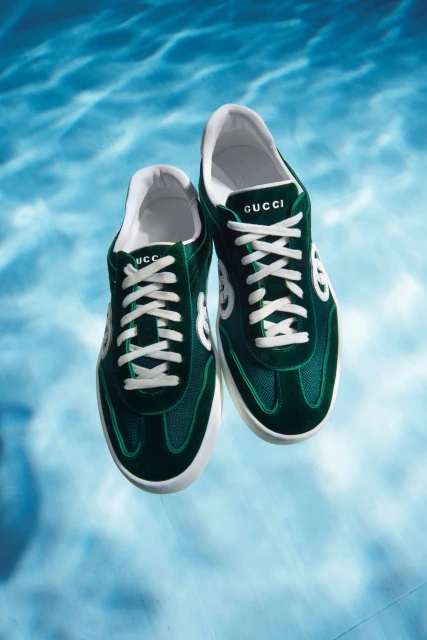 A pair of green Gucci sneakers floating in a blue water-like background, showcased in an eye-level studio product shot.