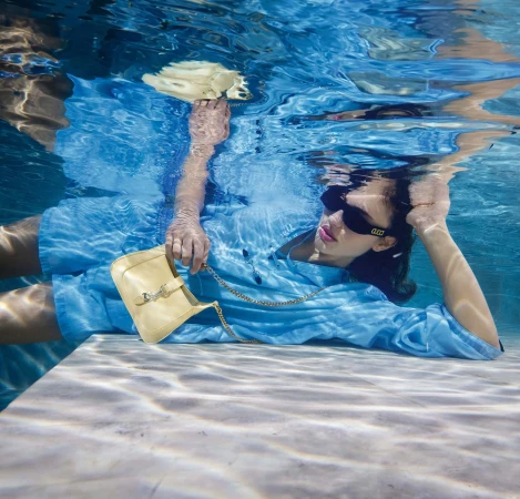 Model wearing blue clothing underwater holding a yellow handbag while lying on a submerged ledge