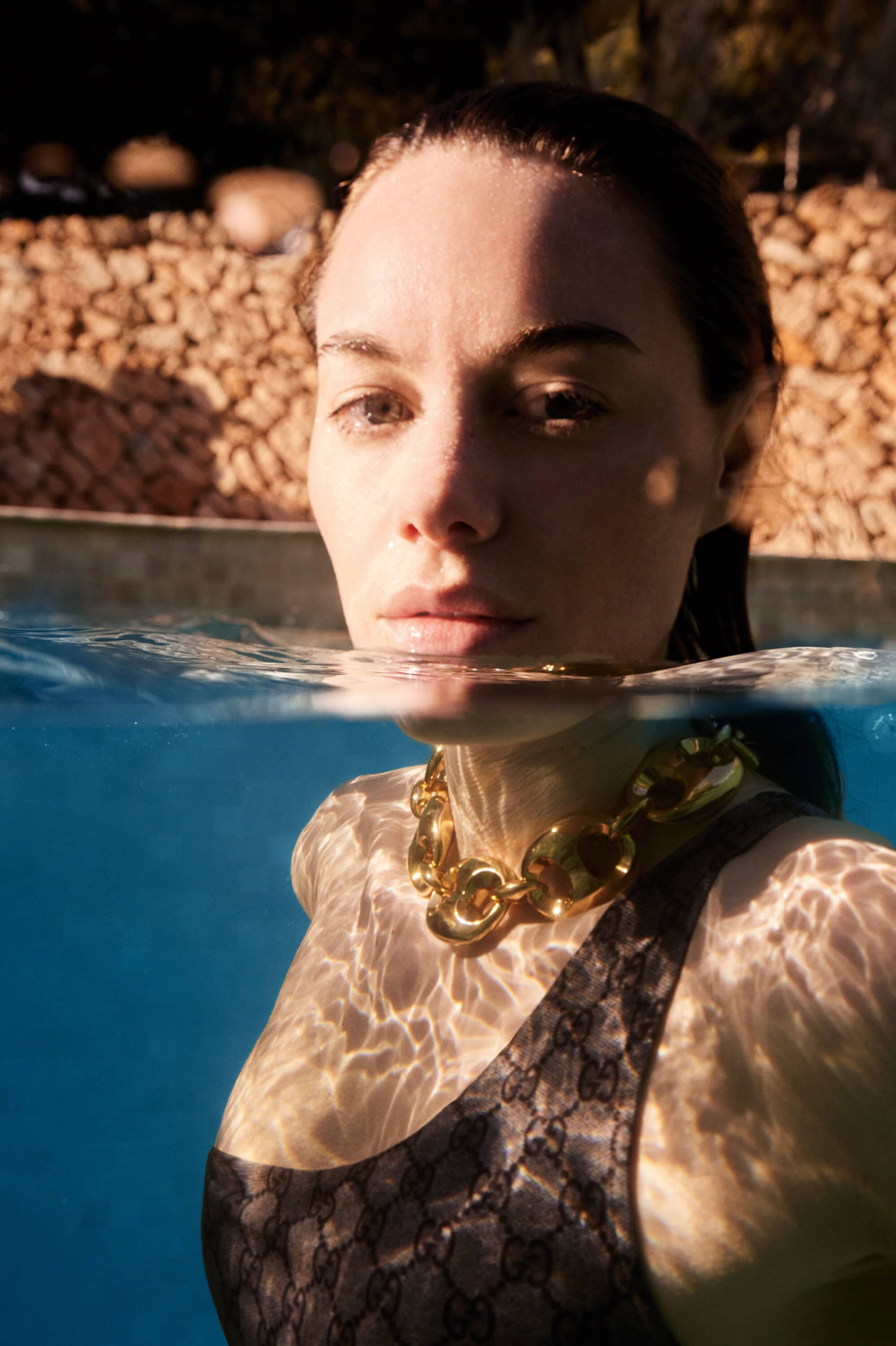 Close-up of a woman in a swimming pool wearing a black swimsuit and gold necklace, water reflects sunlight, creating a bright, summery mood.