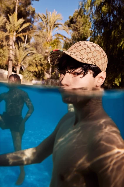 Man with Gucci cap swims in pool, half-submerged view with clear blue water, palm trees, and another person visible in background.