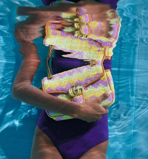 Fashion shot of a person in a purple swimsuit holding a yellow and purple Gucci handbag while submerged in a pool of water.