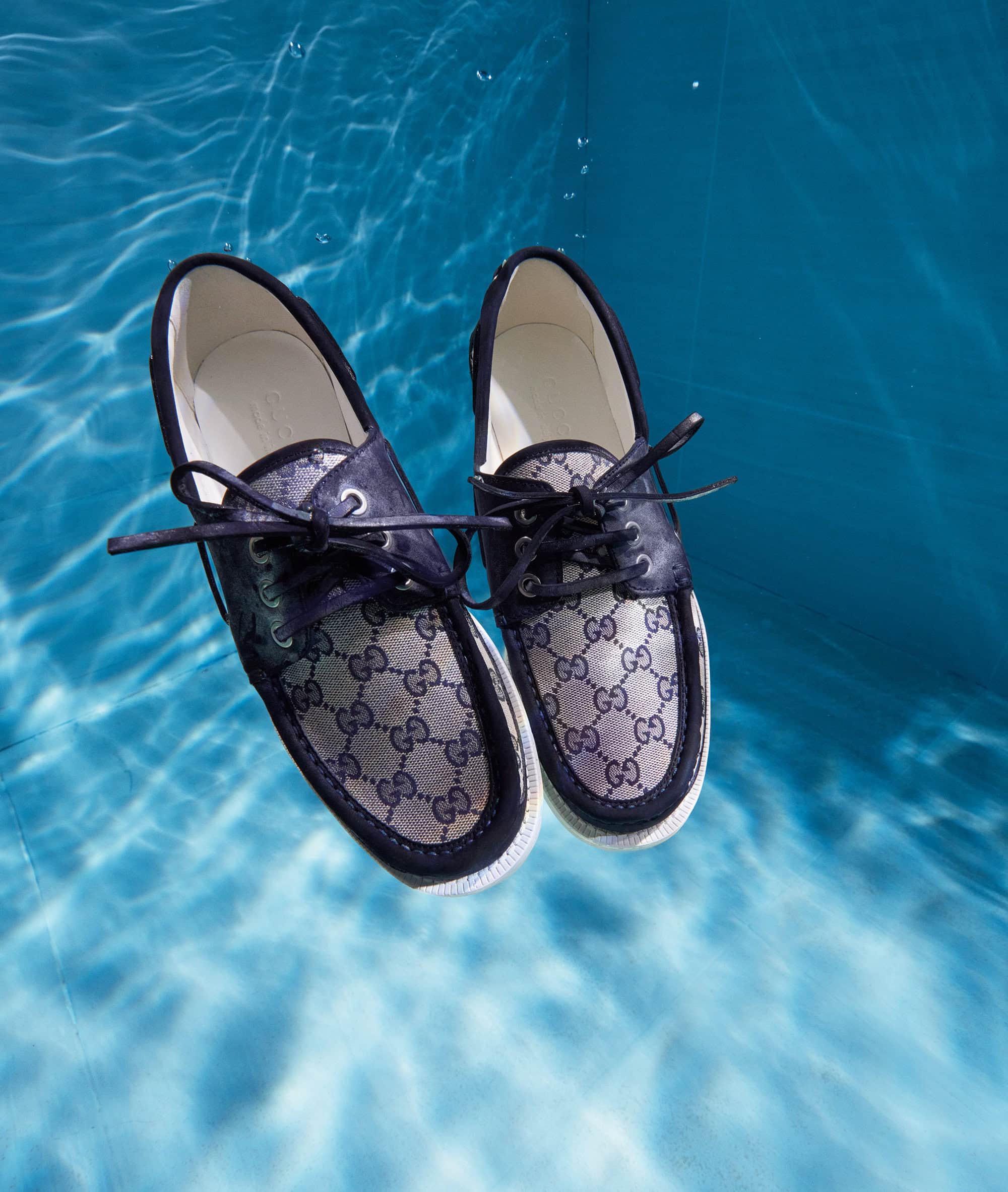 Two Gucci boat shoes float in clear blue water with light reflections, creating an unusual and visually striking composition.