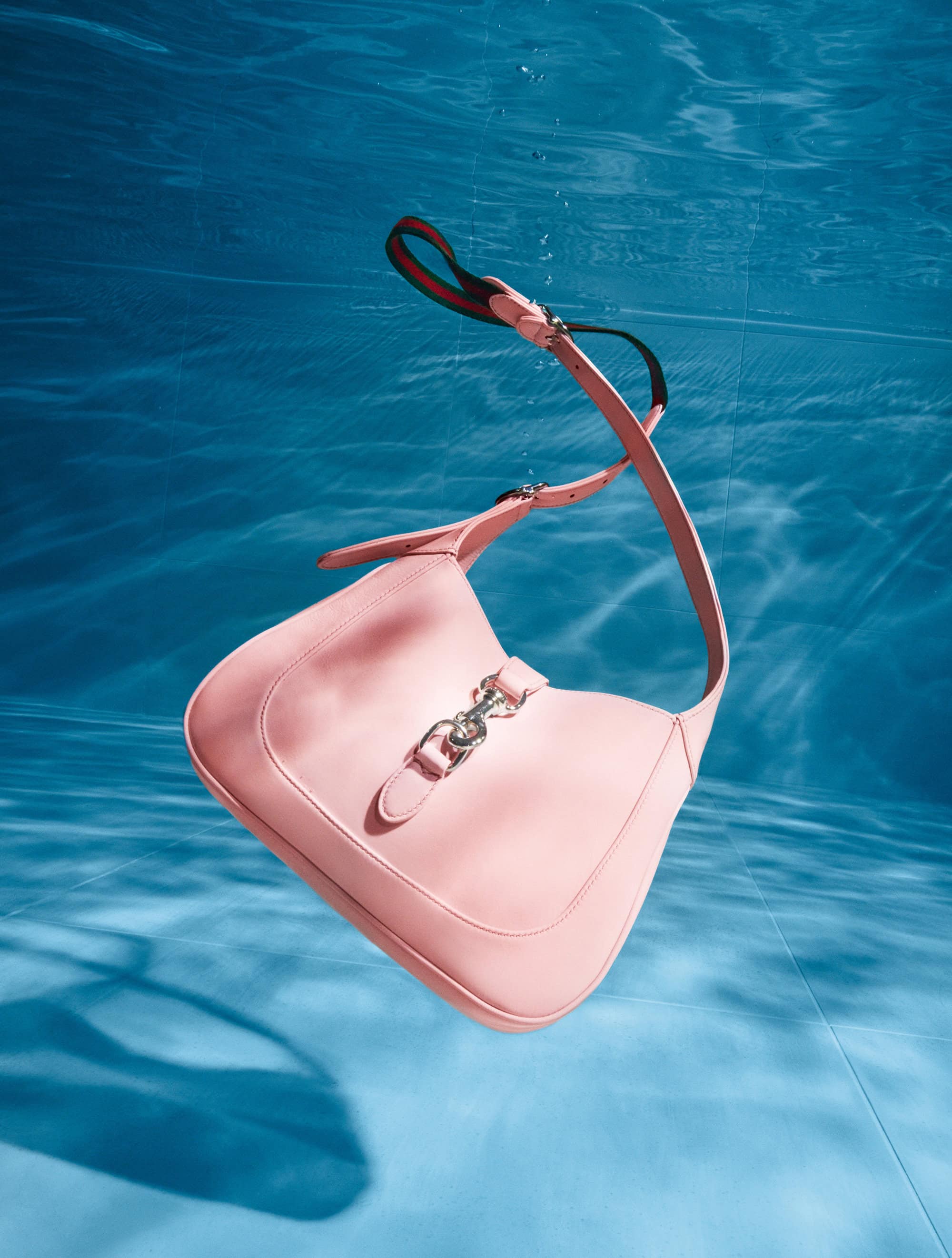 Pale pink leather shoulder bag with silver hardware floats underwater against a mottled blue background with light patterns.