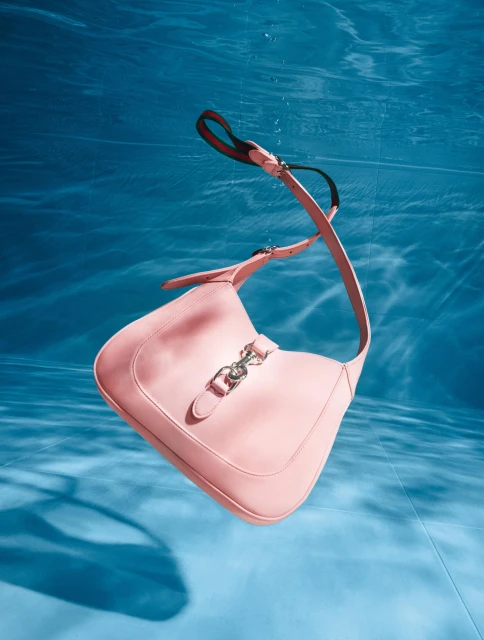 Pale pink leather shoulder bag with silver hardware floats underwater against a mottled blue background with light patterns.