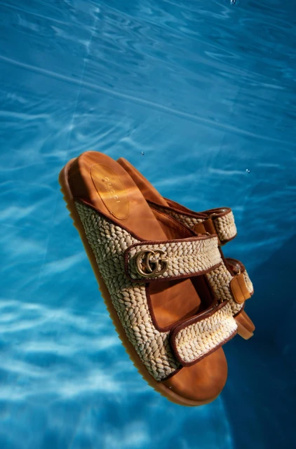 High-angle shot of a pair of sandal slides with beige woven straps and brown soles floating underwater in a swimming pool.