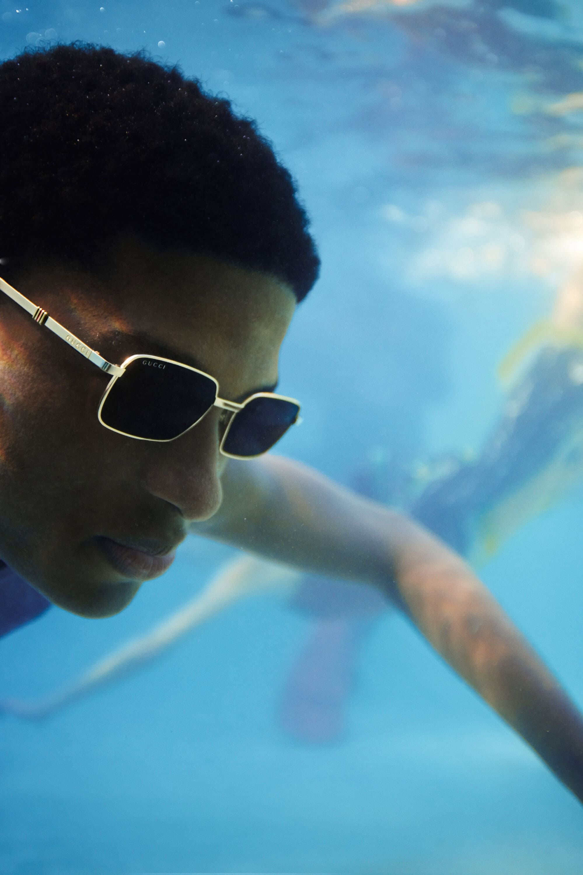 Underwater portrait of a person wearing Gucci sunglasses, with other people blurred in the background, and underwater sun rays visible.