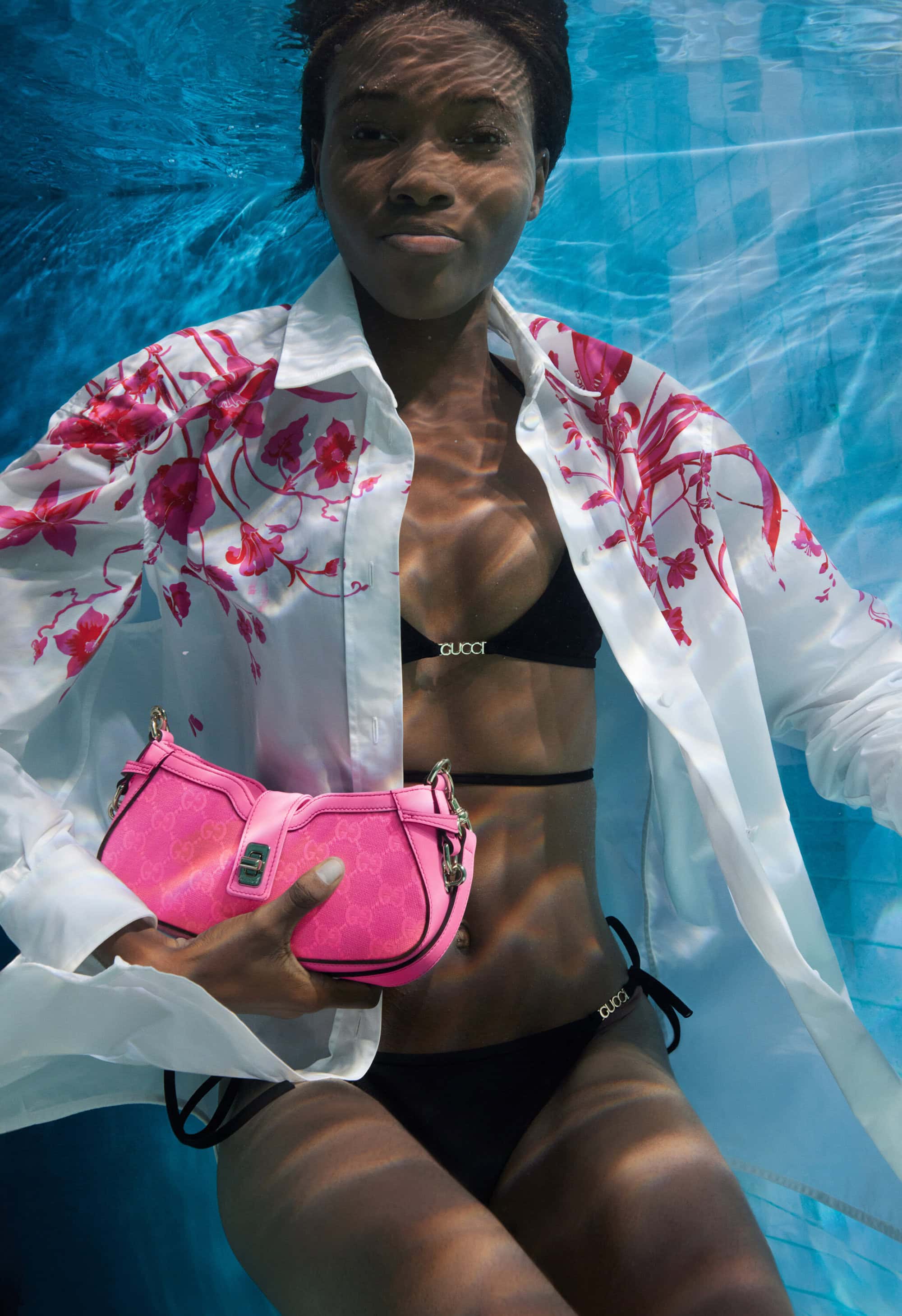 Underwater fashion shot of a woman in a black bikini and floral shirt, holding a pink purse, in a blue pool.