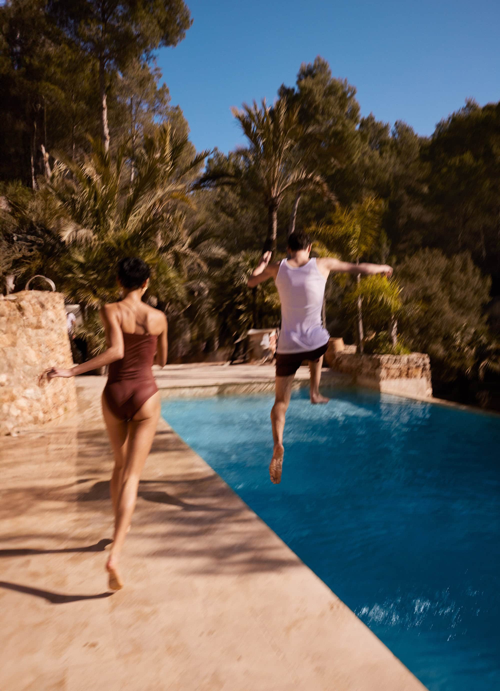 Woman runs toward a pool as a man jumps in, surrounded by trees on a sunny day. The scene conveys summer fun and active lifestyle.