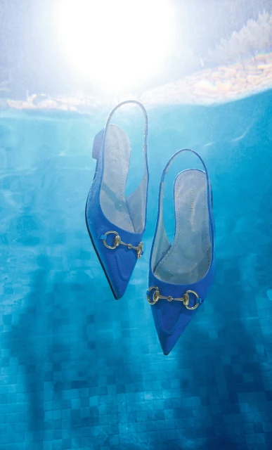 A pair of blue, slingback, pointed-toe high heel shoes with gold hardware floats in clear blue water under bright, diffused sunlight.