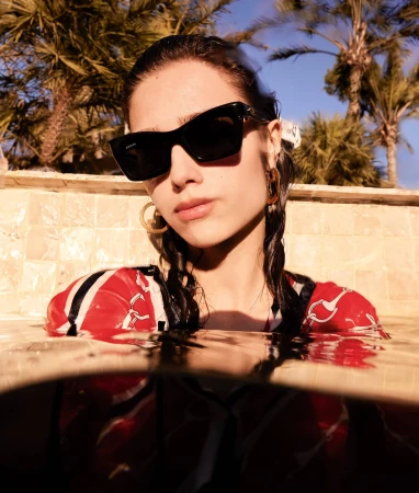 Wet model in a red printed top and sunglasses partially submerged in a pool with palm trees in the background