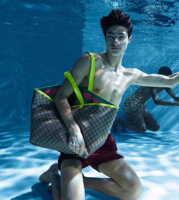 Underwater shot of a young man kneeling in a pool, holding a Gucci tote bag with neon yellow straps, and another man swimming in the background.