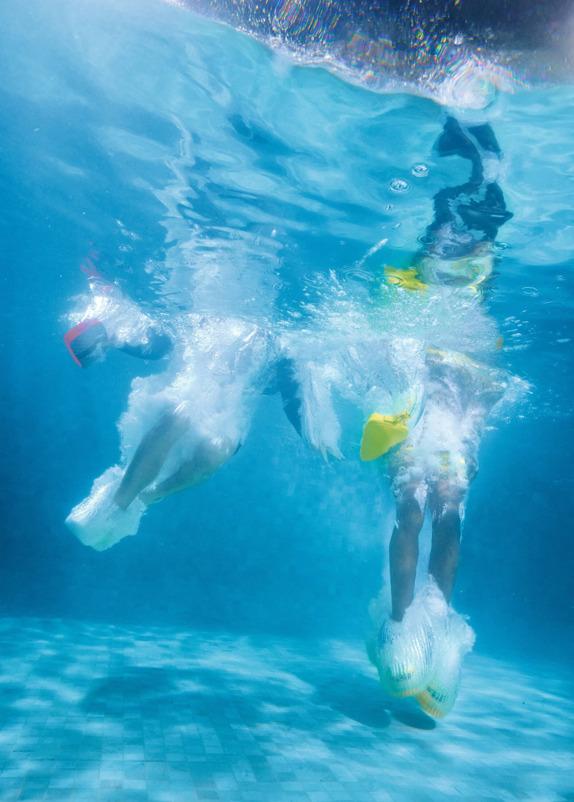 Underwater shot of three people with their legs visible, submerged in a swimming pool with light blue water and tile flooring.