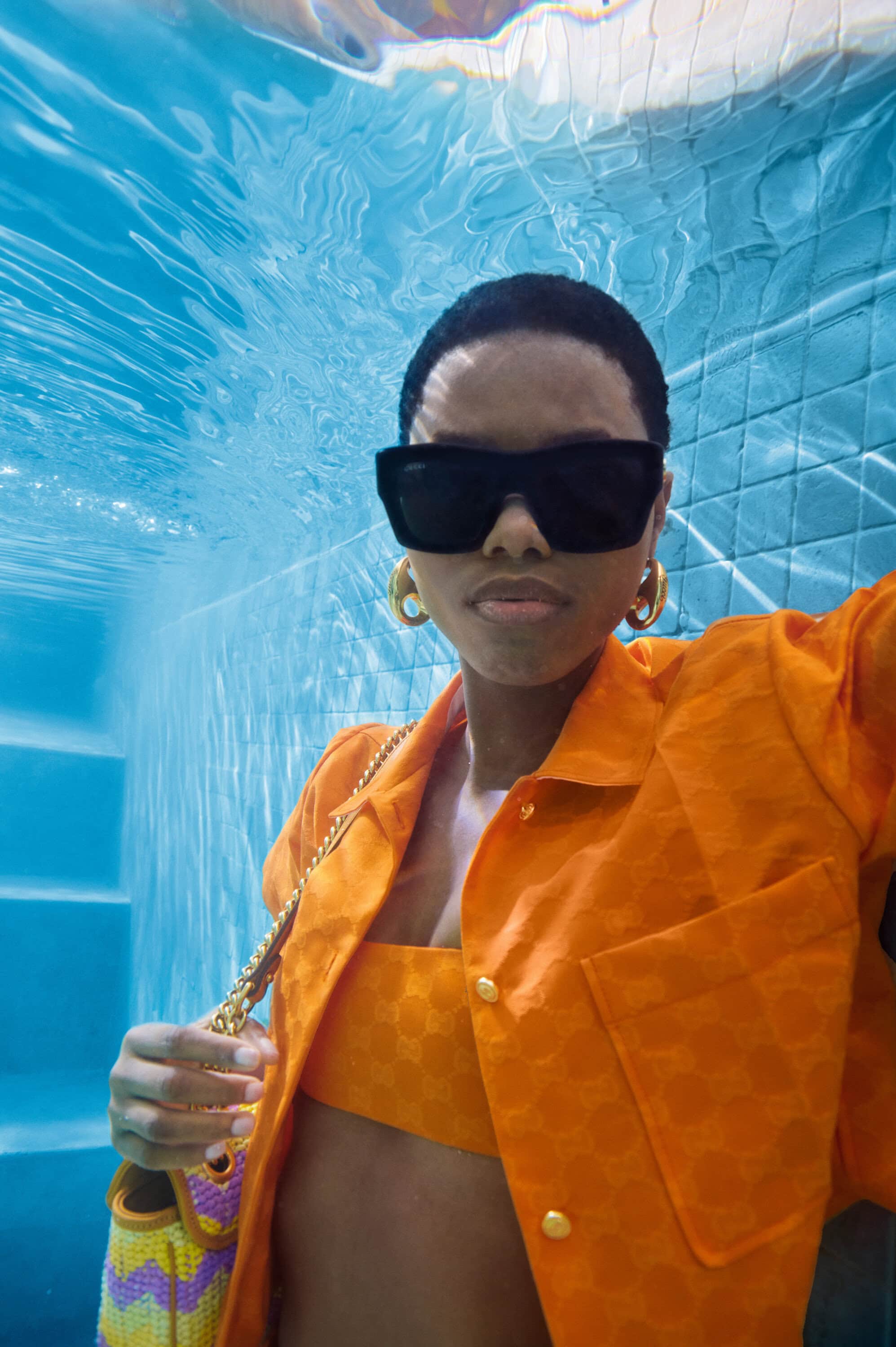 Underwater selfie of a woman with sunglasses and an orange outfit. She is wearing hoop earrings and holding a purse.