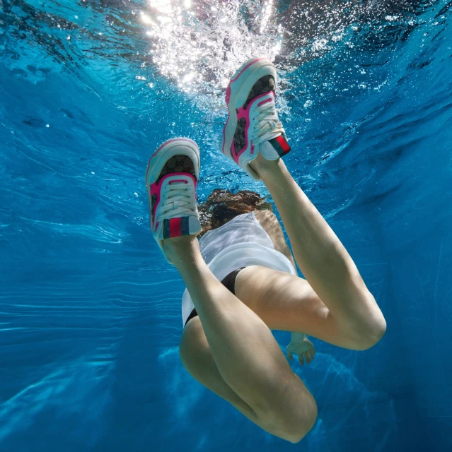 Underwater view of a woman swimming in a pool, wearing sneakers, captured with bright lighting and clear water.